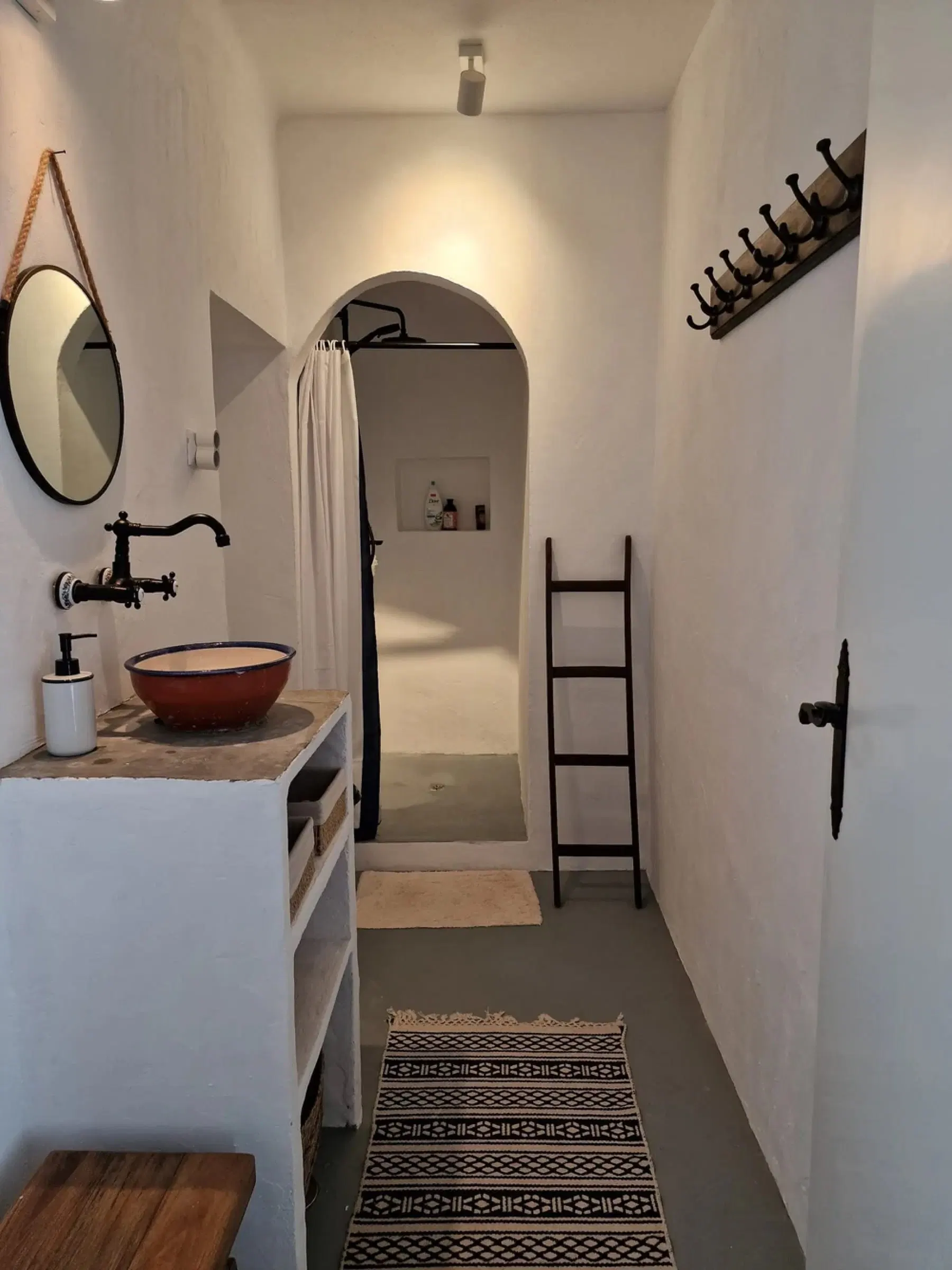 A whitewashed bathroom with a clay basin on a stone counter, a round mirror and a shower set in an arched alcove.