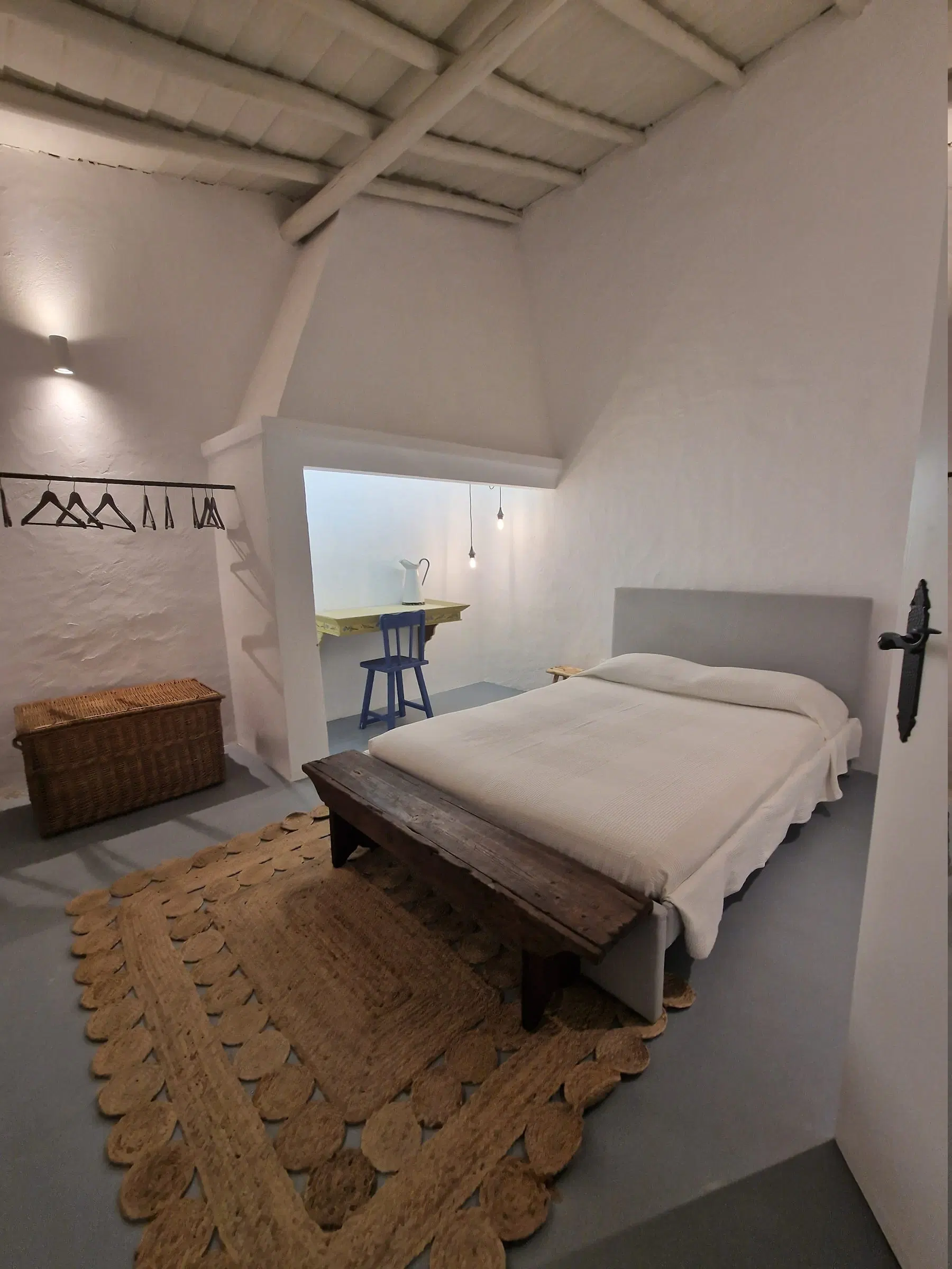 A single bed in a whitewashed room, a wicker chest at its foot and an arched doorway beyond.