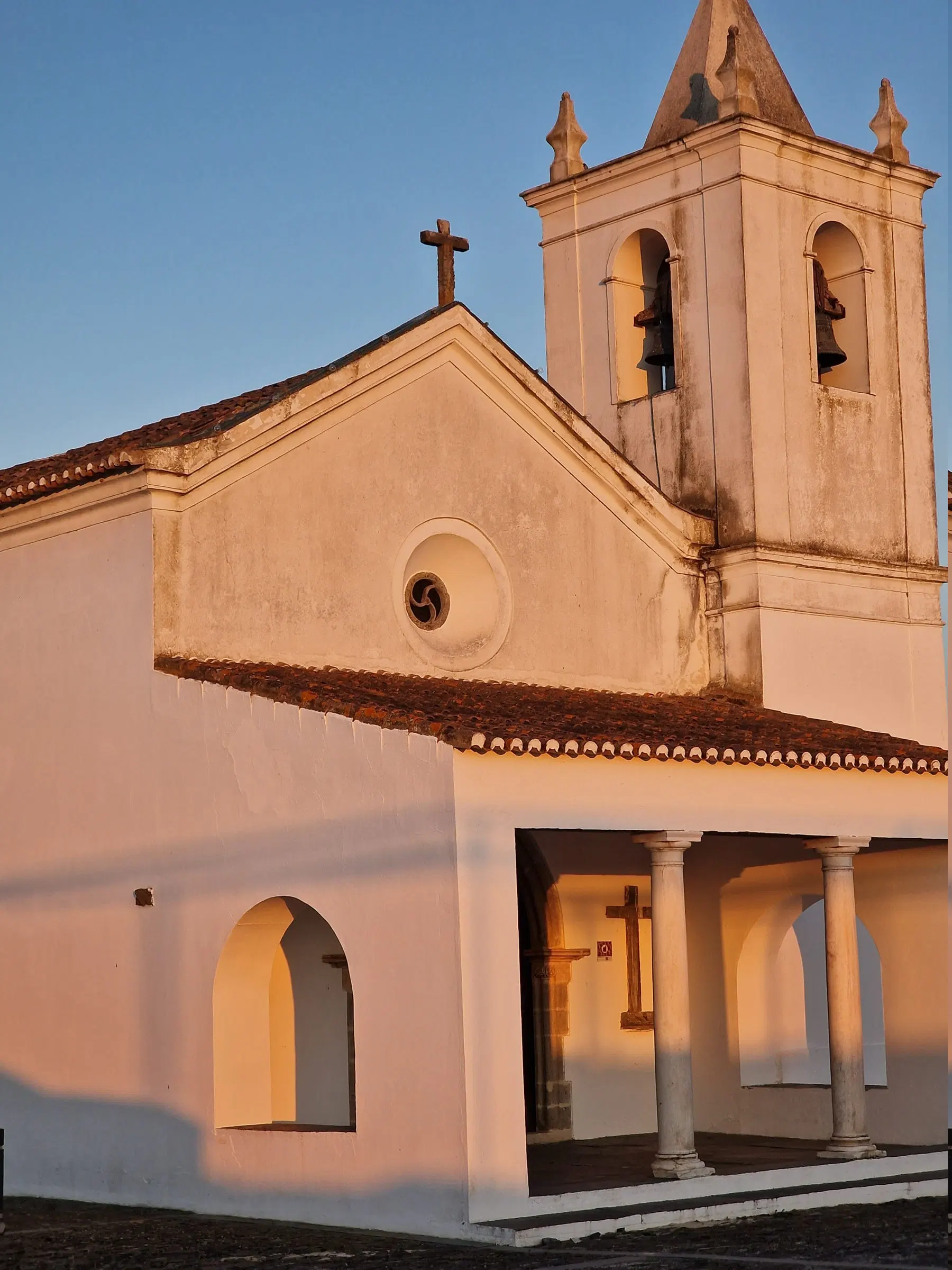 A igreja da Aldeia da Luz, caiada, com alpendre de arcos e cunhais em pedra, na última luz do dia.