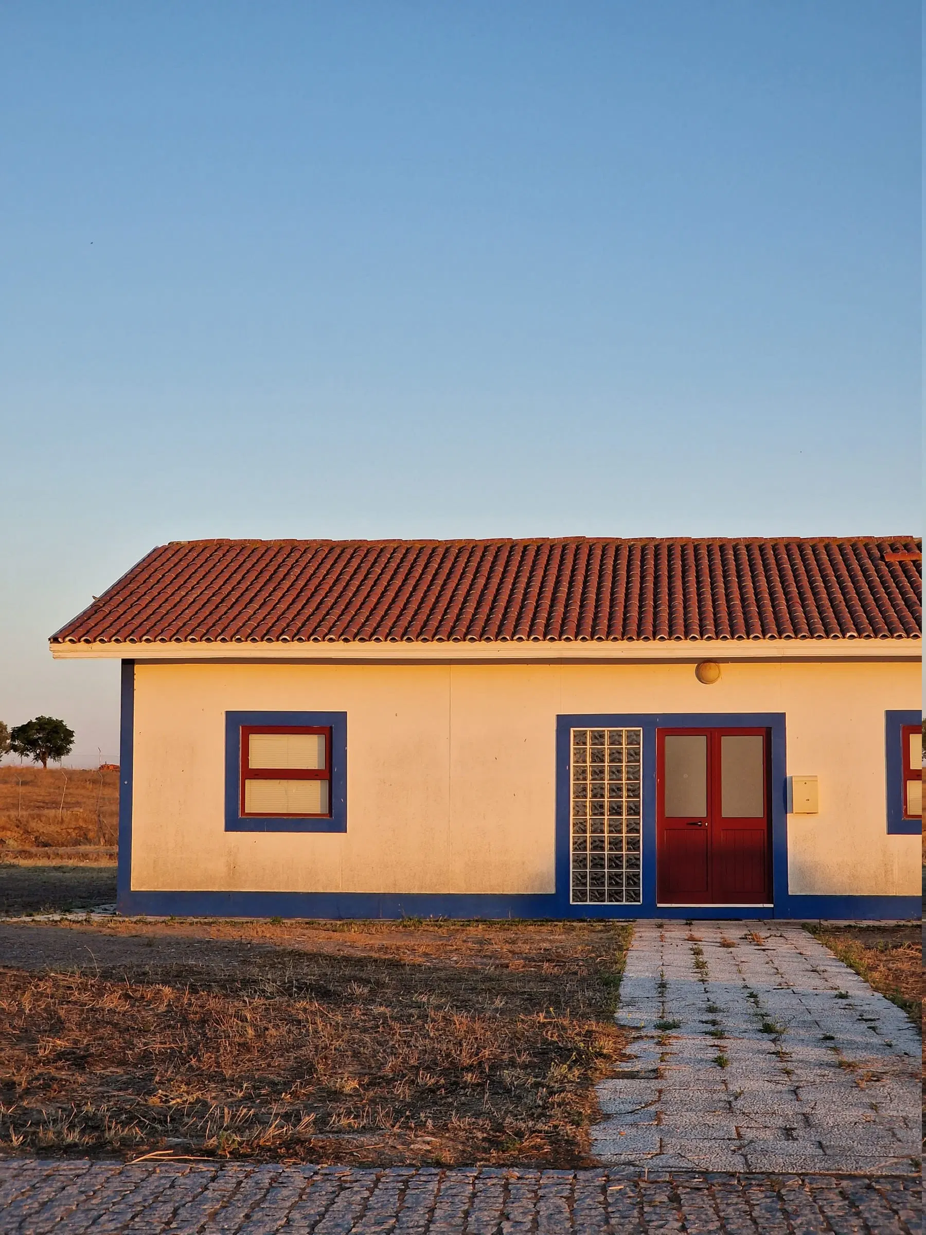 Edifício baixo caiado, de portas vermelhas e barras azuis, sozinho na planície.