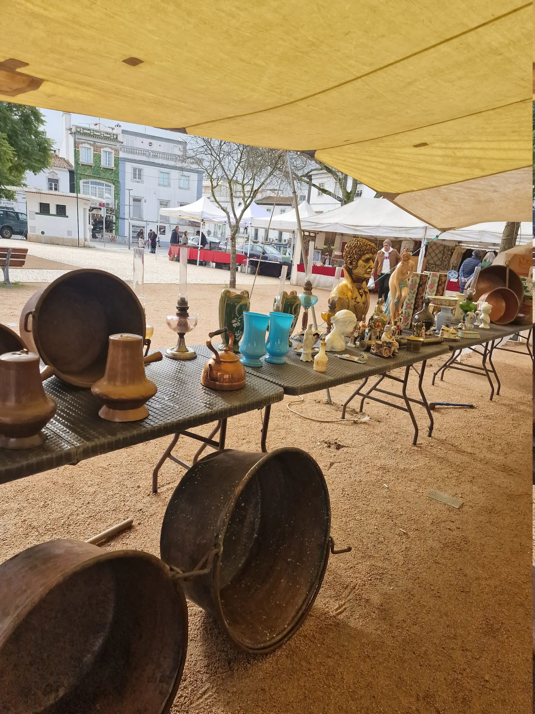 Mesa de feira de velharias, com peças de cobre, louça e candeeiros antigos.