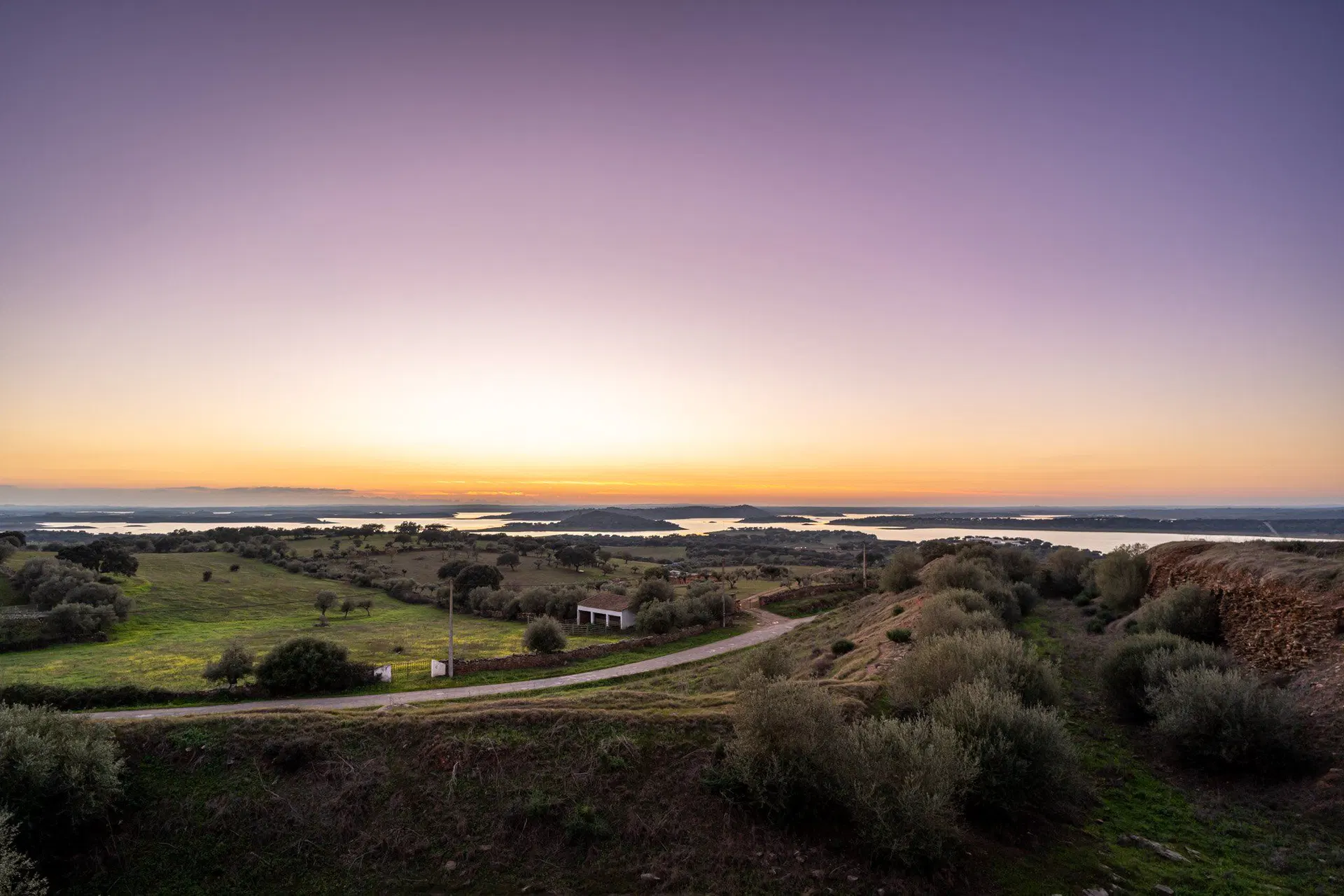 O Grande Lago do Alqueva ao anoitecer, com os braços de água a perder-se entre montados e um caminho de terra a descer para a albufeira.
