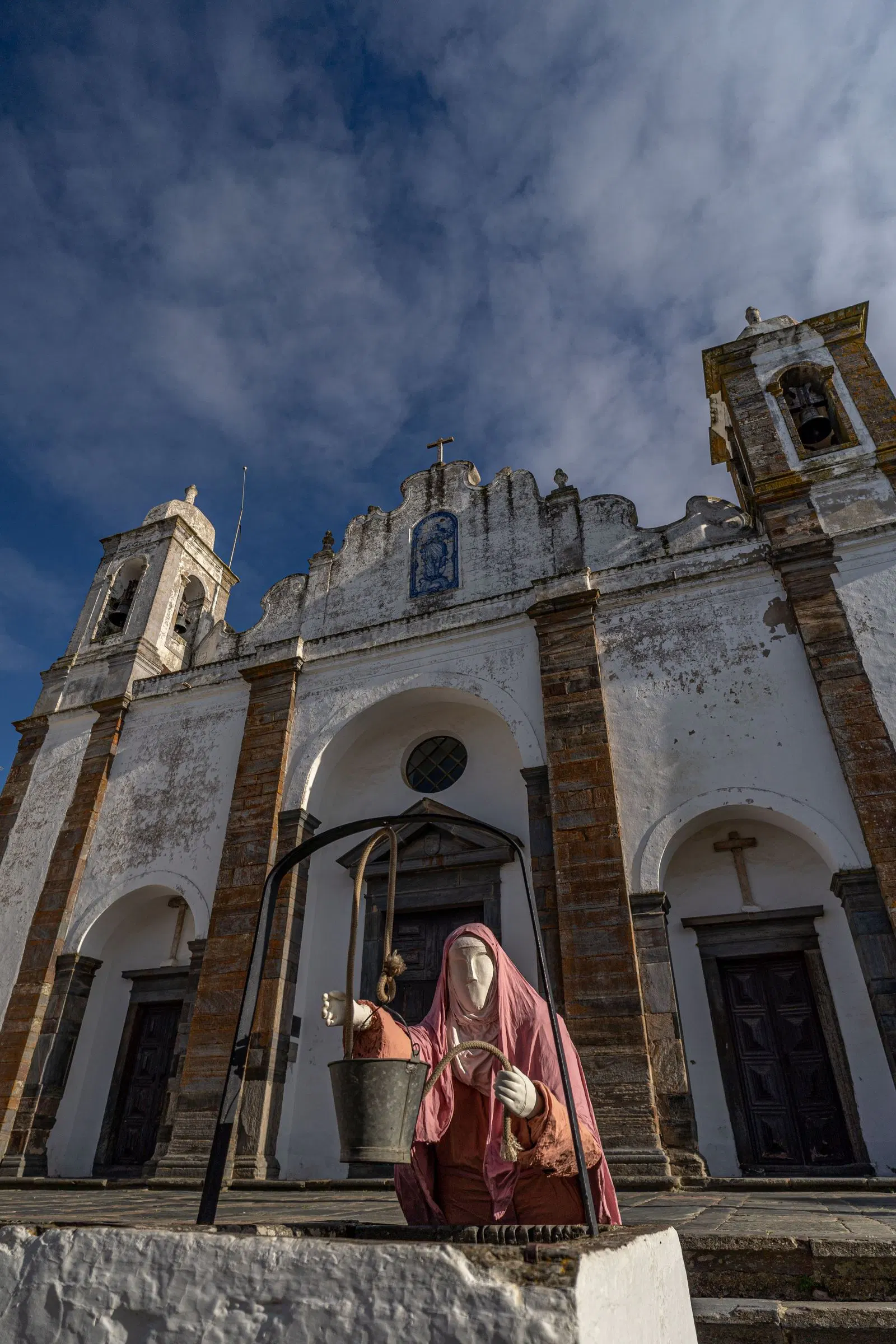 La fachada blanca y ocre de la iglesia parroquial de Monsaraz, con sus dos campanarios, y una figura con traje de época frente a la picota.