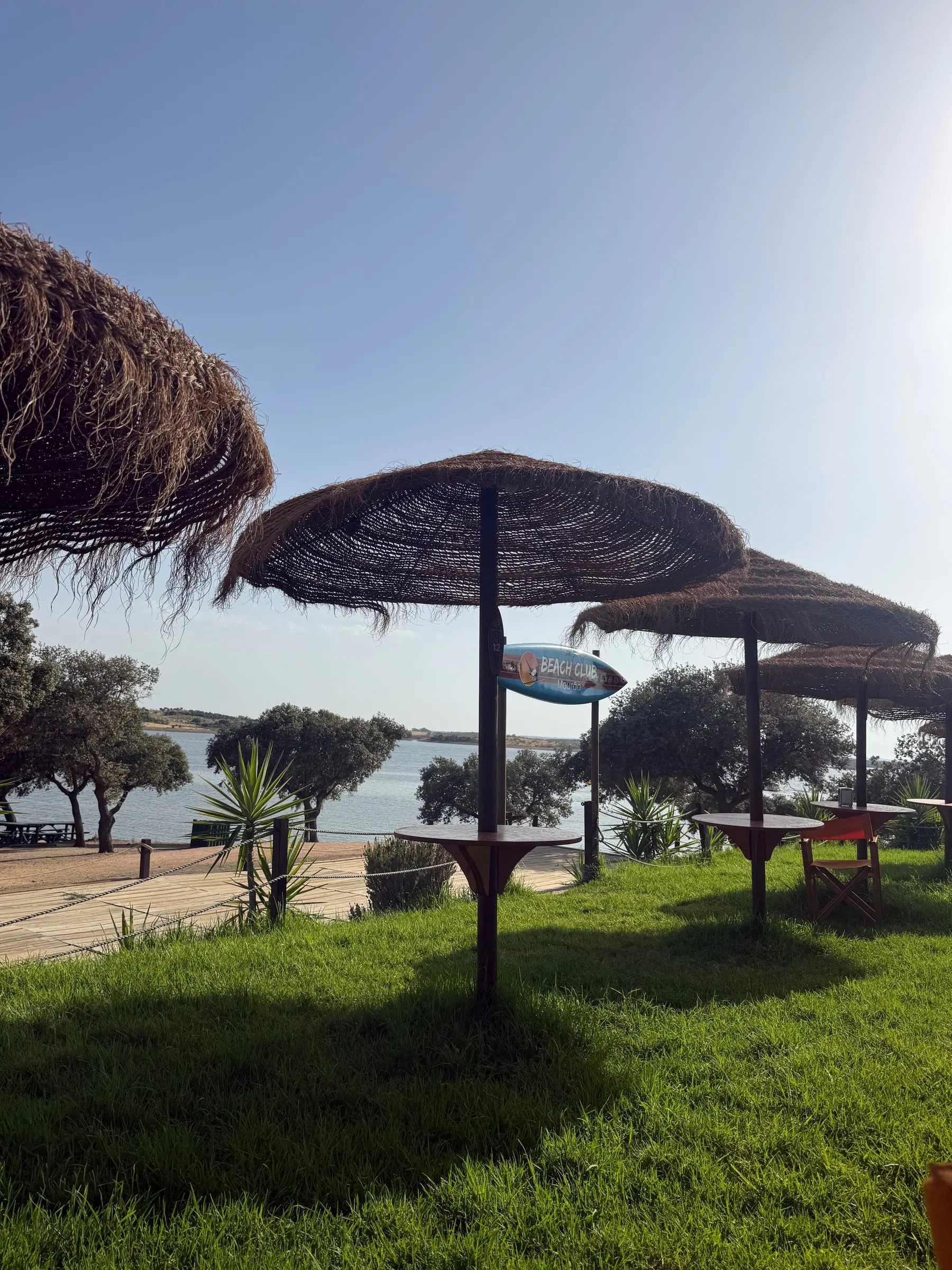 Césped, sombrillas y mesas junto al Beach Club de la Playa Fluvial de Mourão, con el lago al fondo.