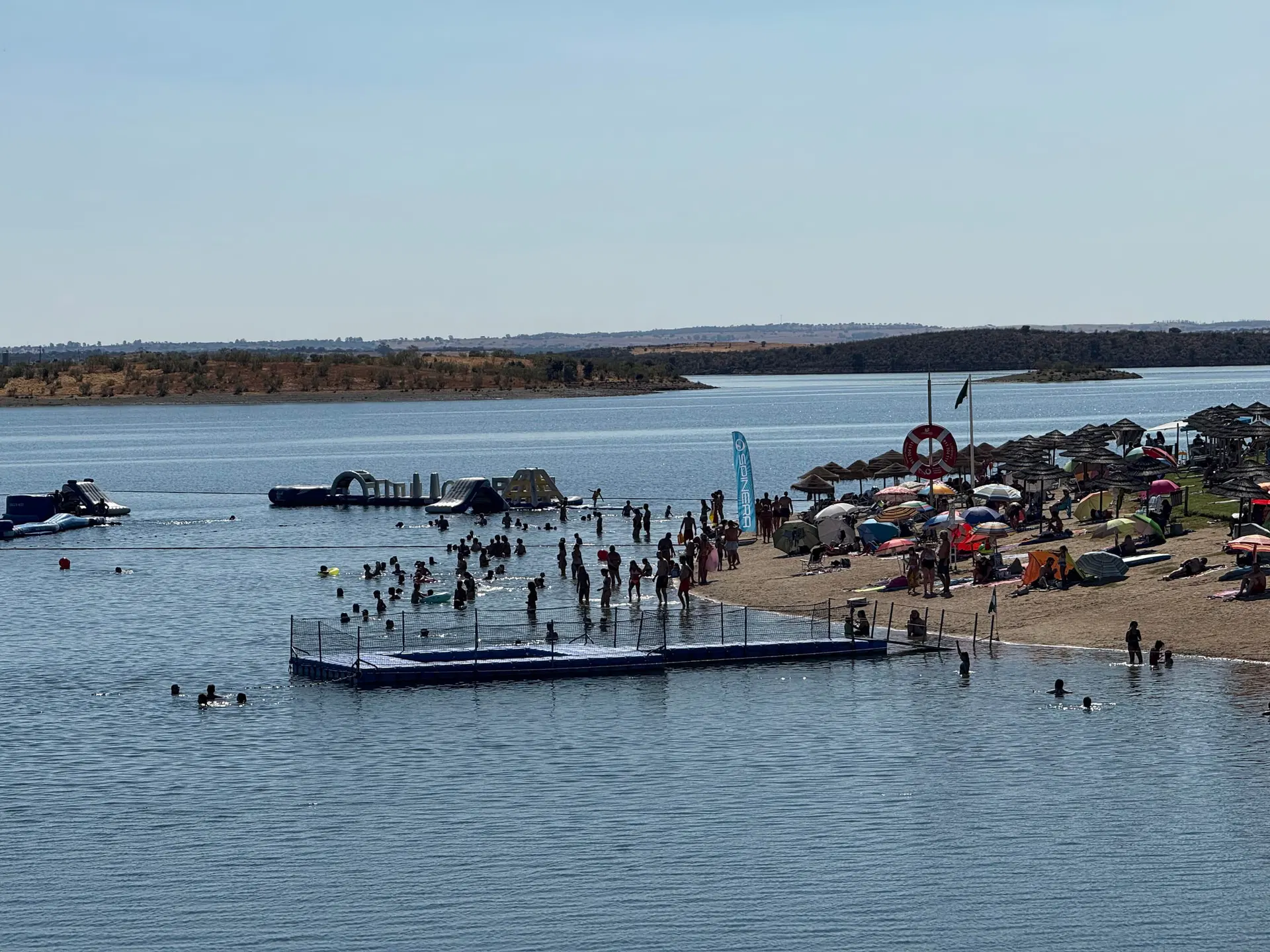 A Praia Fluvial de Mourão num dia de verão, com banhistas, piscina flutuante, areal e chapéus de sol junto ao Alqueva.