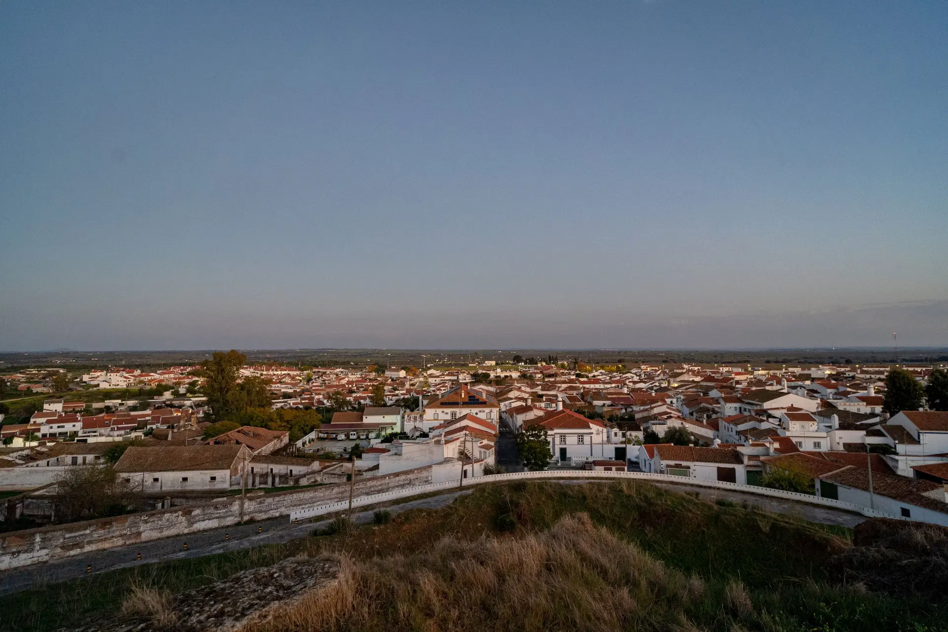 A vila de Mourão vista de cima ao fim da tarde: telhados de telha, casas caiadas e a planície a estender-se até ao horizonte.