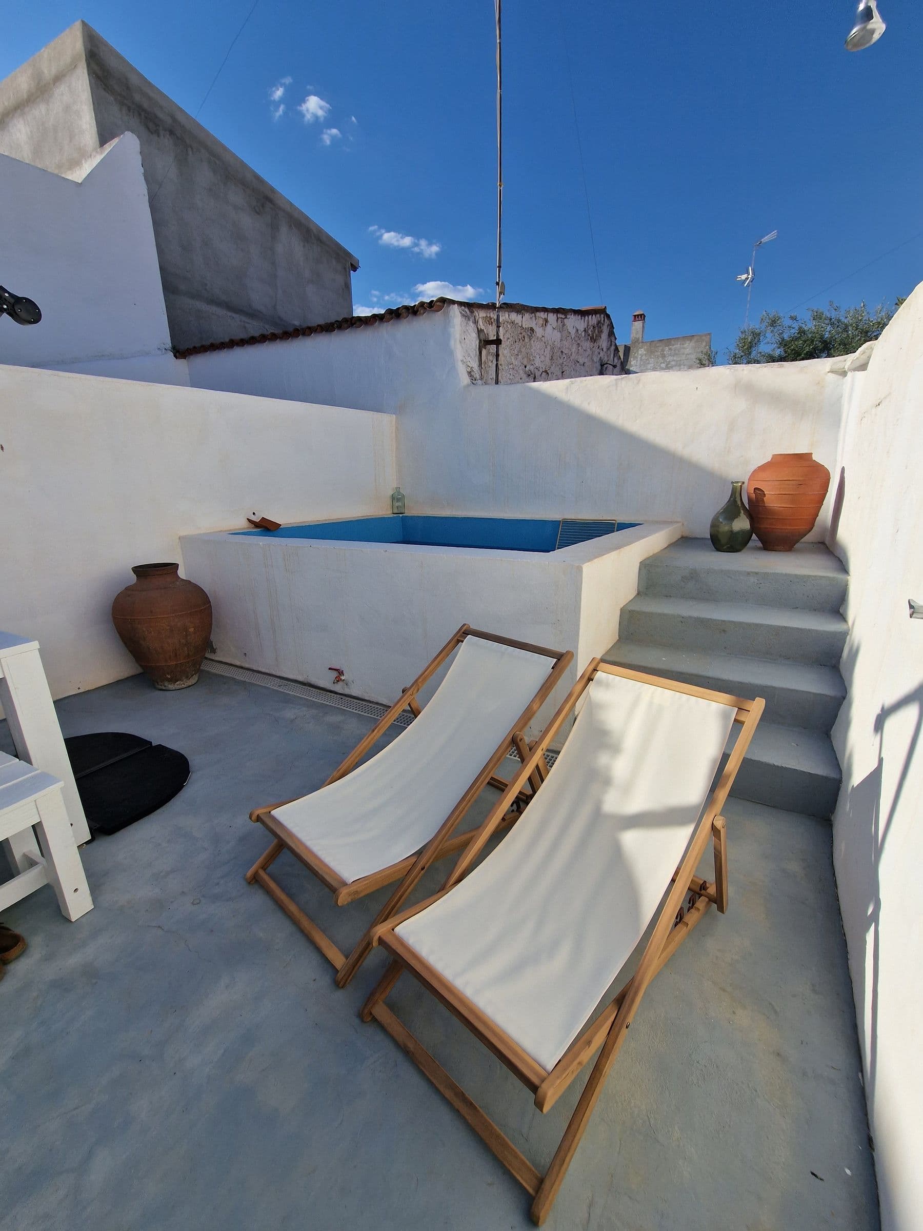 Rectangular plunge pool set into the whitewashed courtyard, with two white canvas and wood deckchairs, a clay urn and concrete steps in full sun.