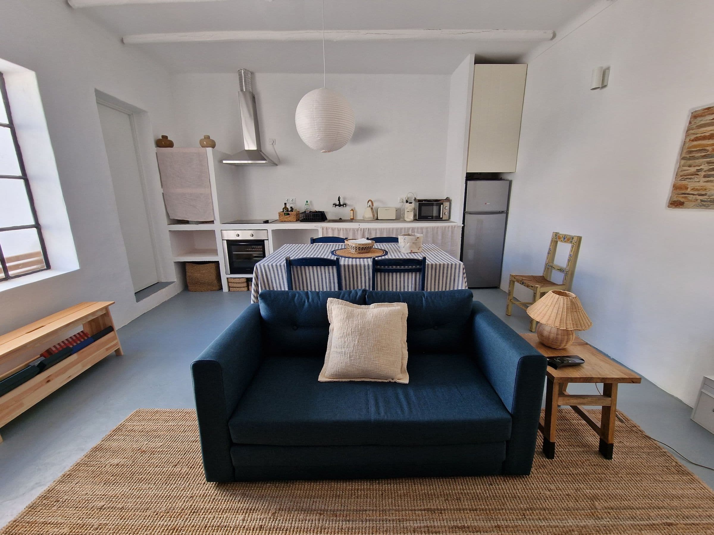 Open-plan living room and kitchen with a deep blue sofa, a table laid with blue-and-white striped linen, a paper lantern and a jute rug.