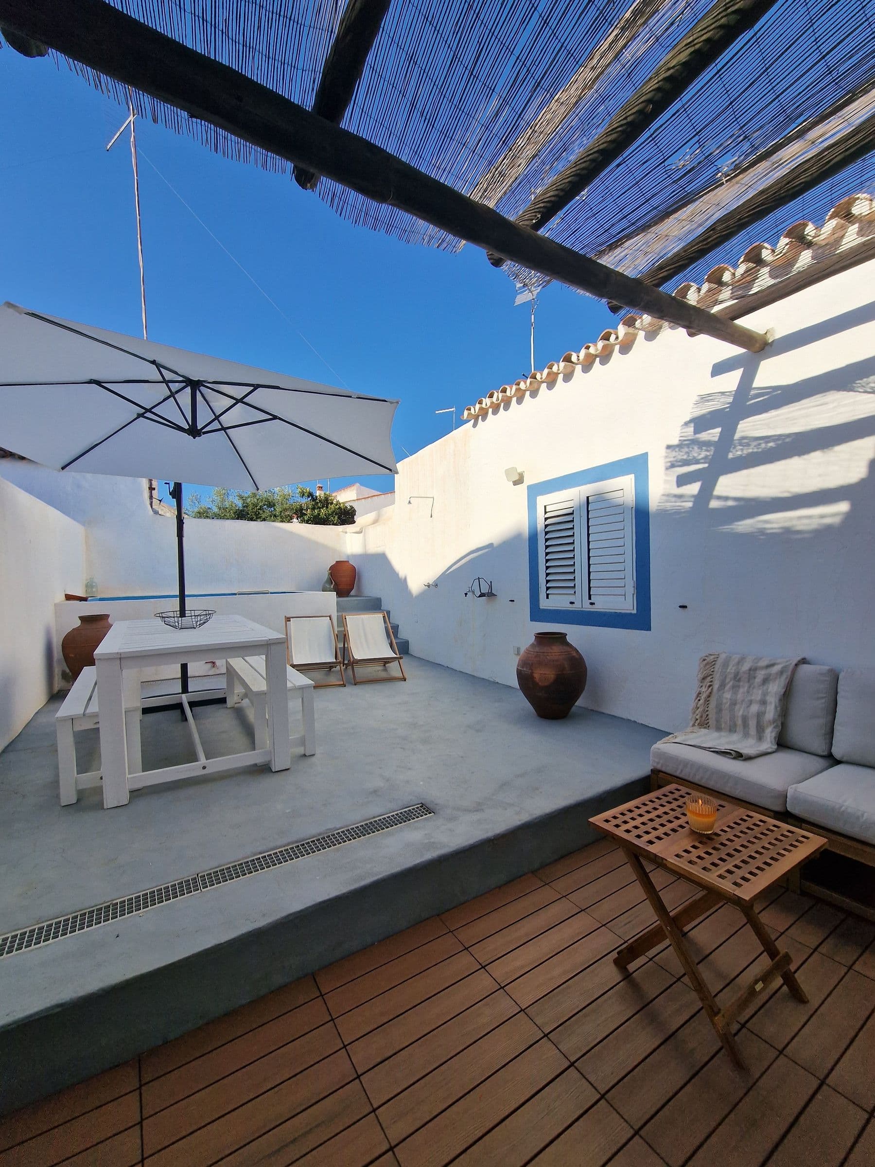 The house terrace with a white table, parasol and built-in sofa shaded by a cane canopy, beside a blue shutter and clay urns.