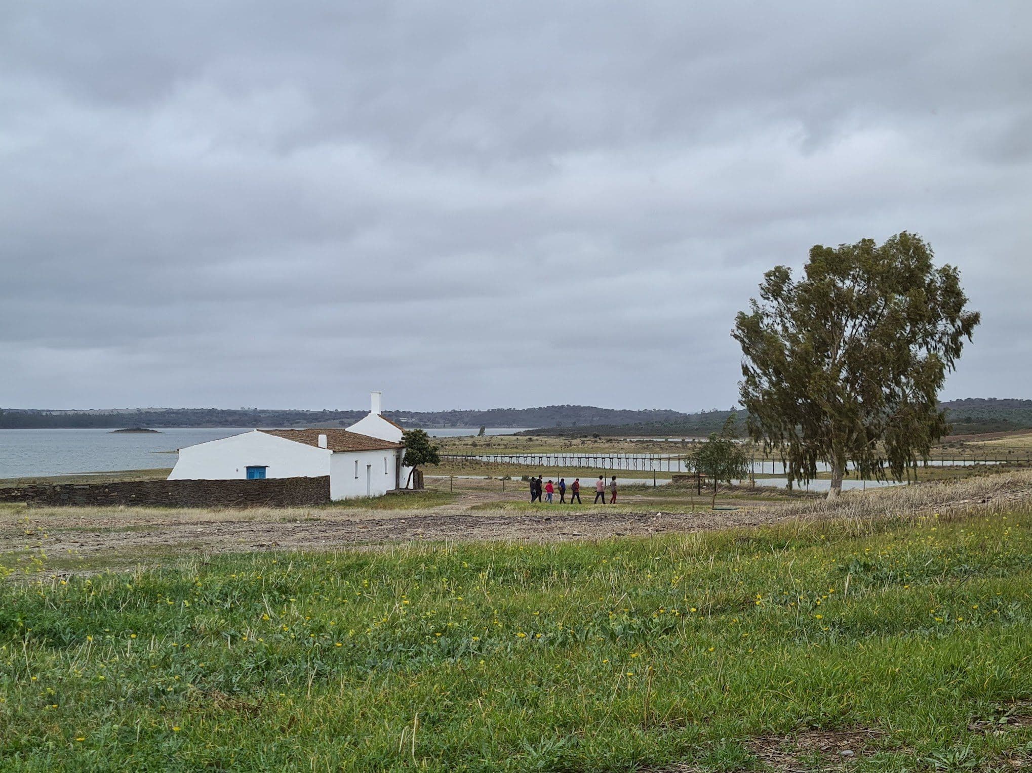 A capela branca da Aldeia da Luz junto à margem do Alqueva, com um grupo de visitantes a caminhar pelo passadiço sob um céu carregado.