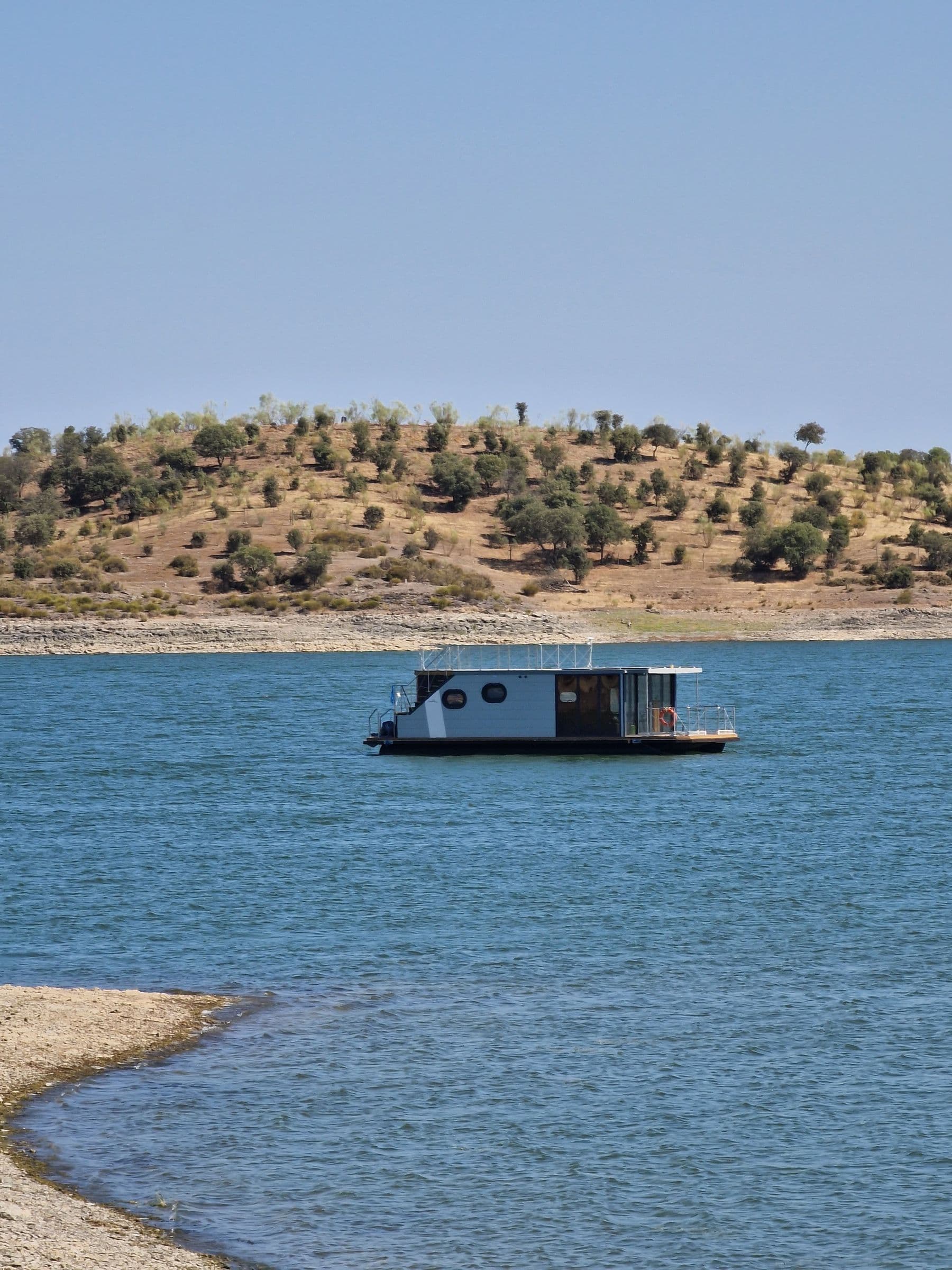 A houseboat moored on the blue water of Alqueva, beside a dry slope dotted with olive trees.