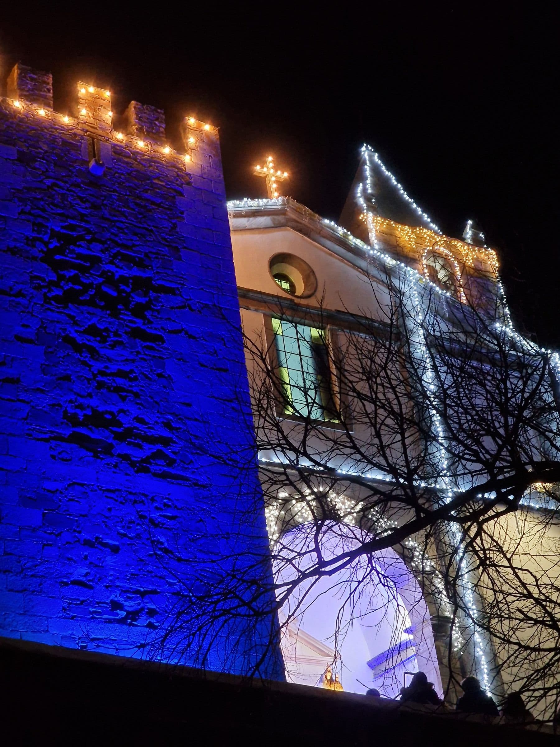 A torre do castelo de Mourão iluminada a azul ao lado da Igreja Matriz decorada com luzes, durante as festas de inverno da vila.