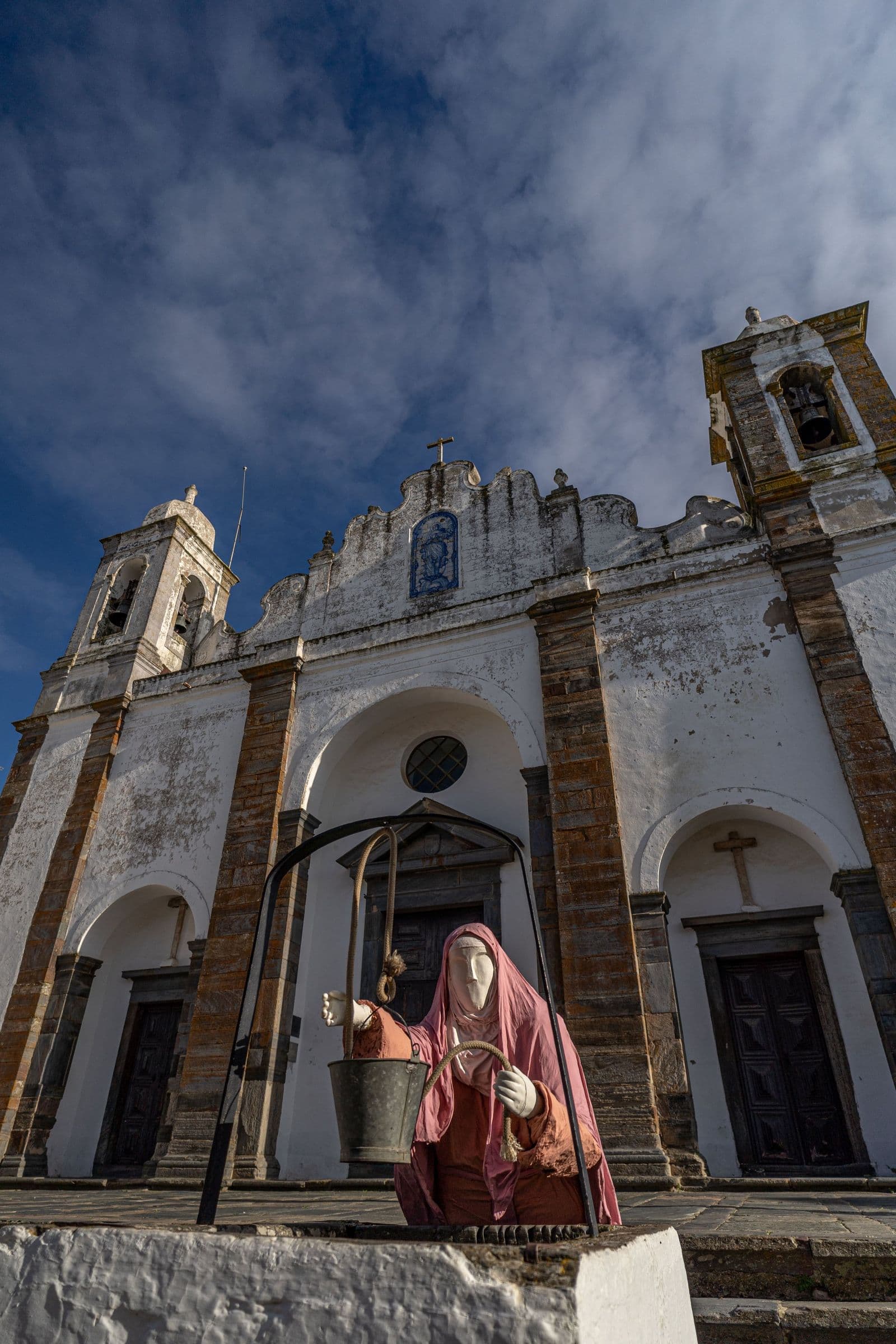 A fachada branca e ocre da Igreja Matriz de Monsaraz, com as suas duas torres sineiras, e uma figura em traje de época em frente ao pelourinho.