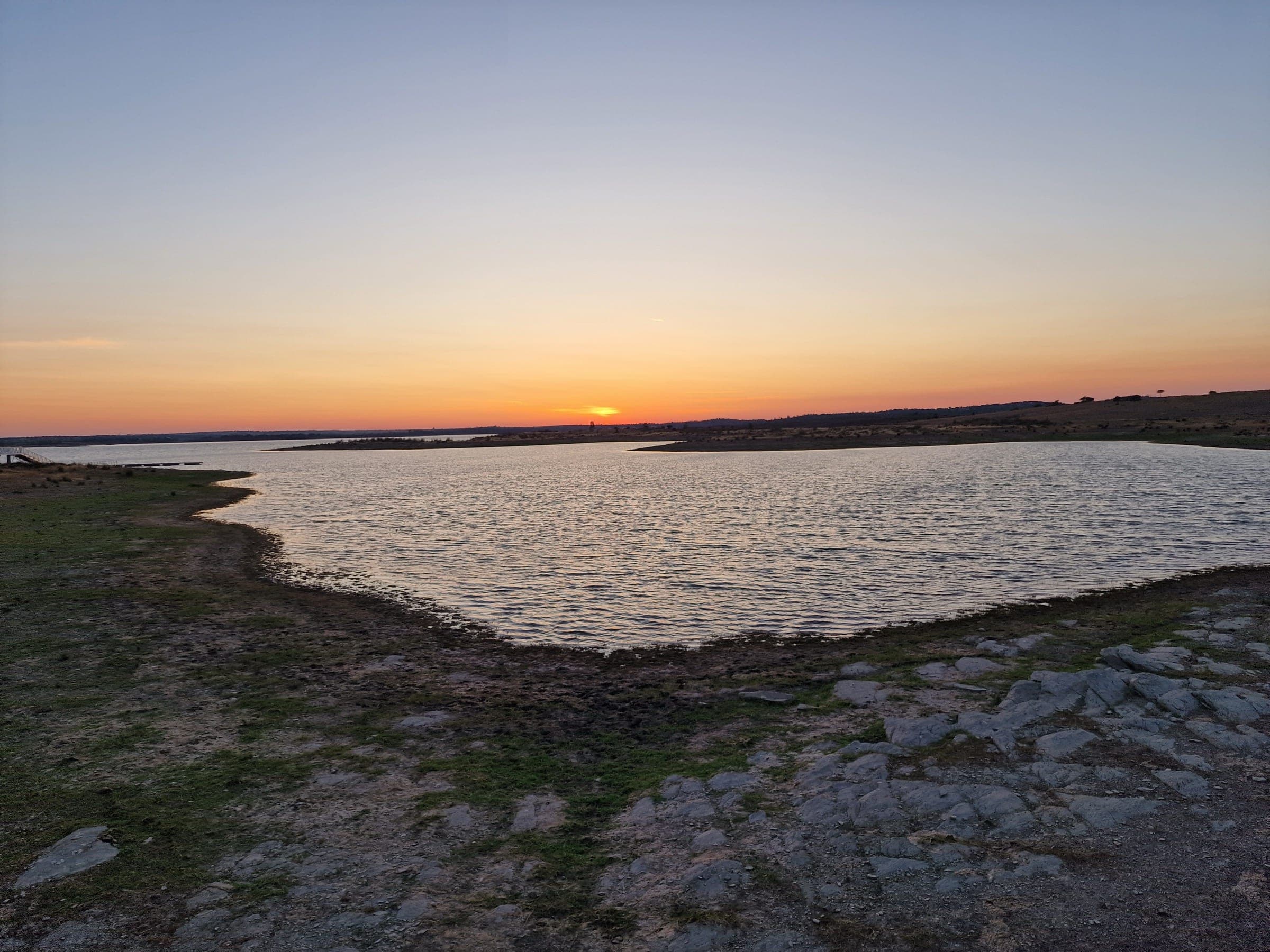 A margem pedregosa da albufeira do Alqueva ao pôr do sol, com a água a perder-se na linha do horizonte.