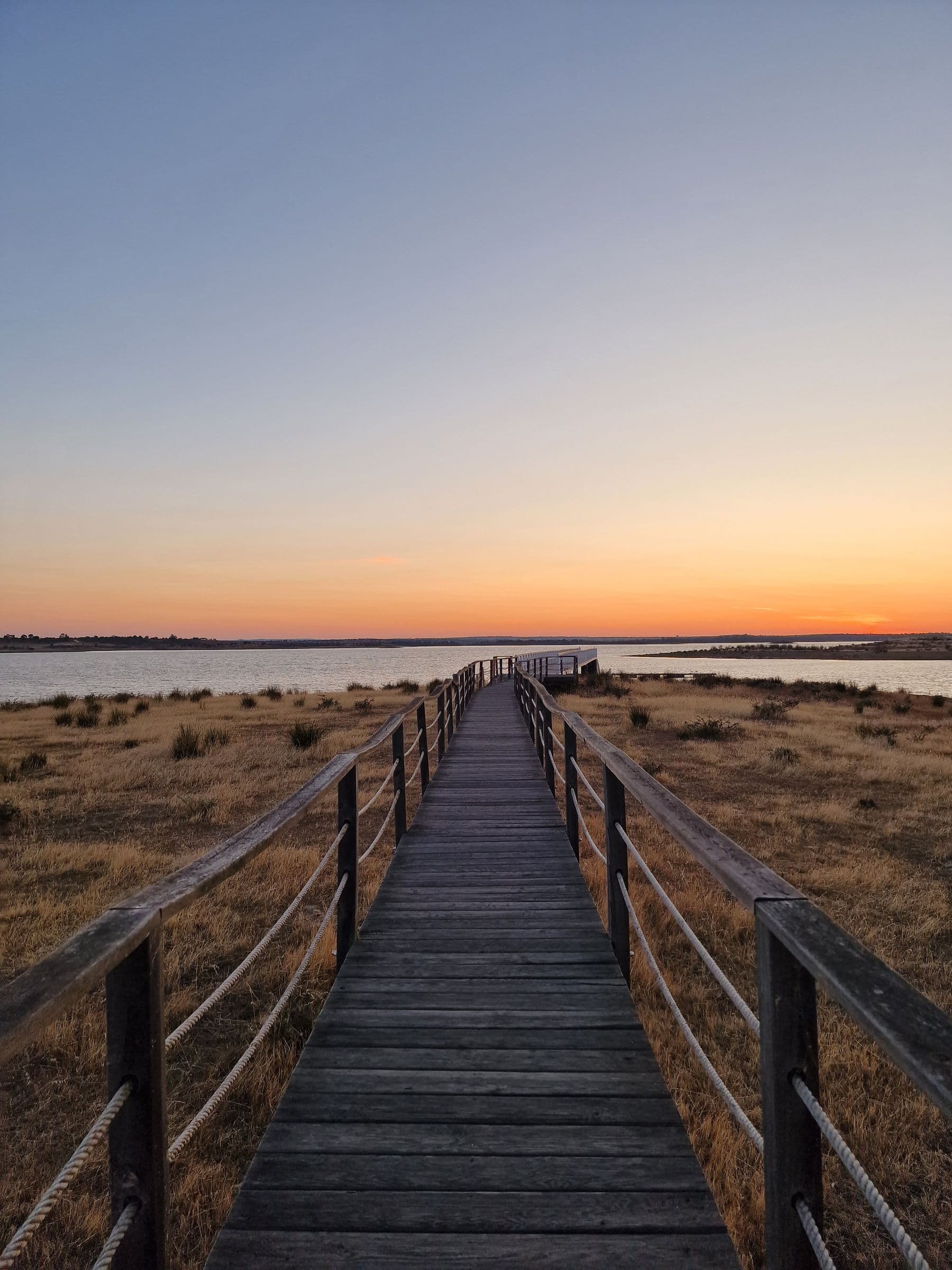 A wooden boardwalk crossing dry scrub towards the water at sunset, on the approach to the Mourão river beach.