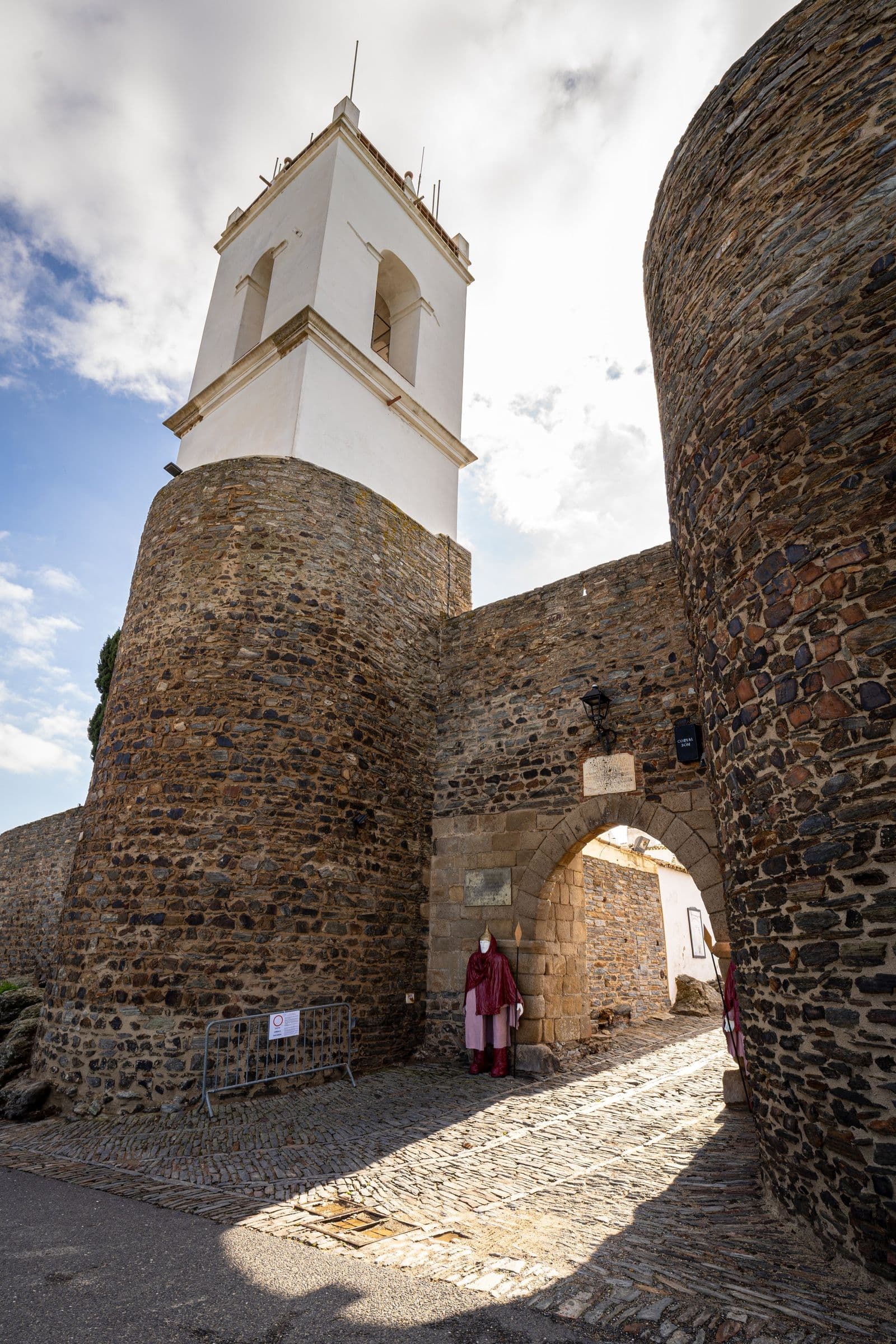 A porta em arco das muralhas de Monsaraz, com a torre branca do relógio a erguer-se sobre a cantaria de xisto.
