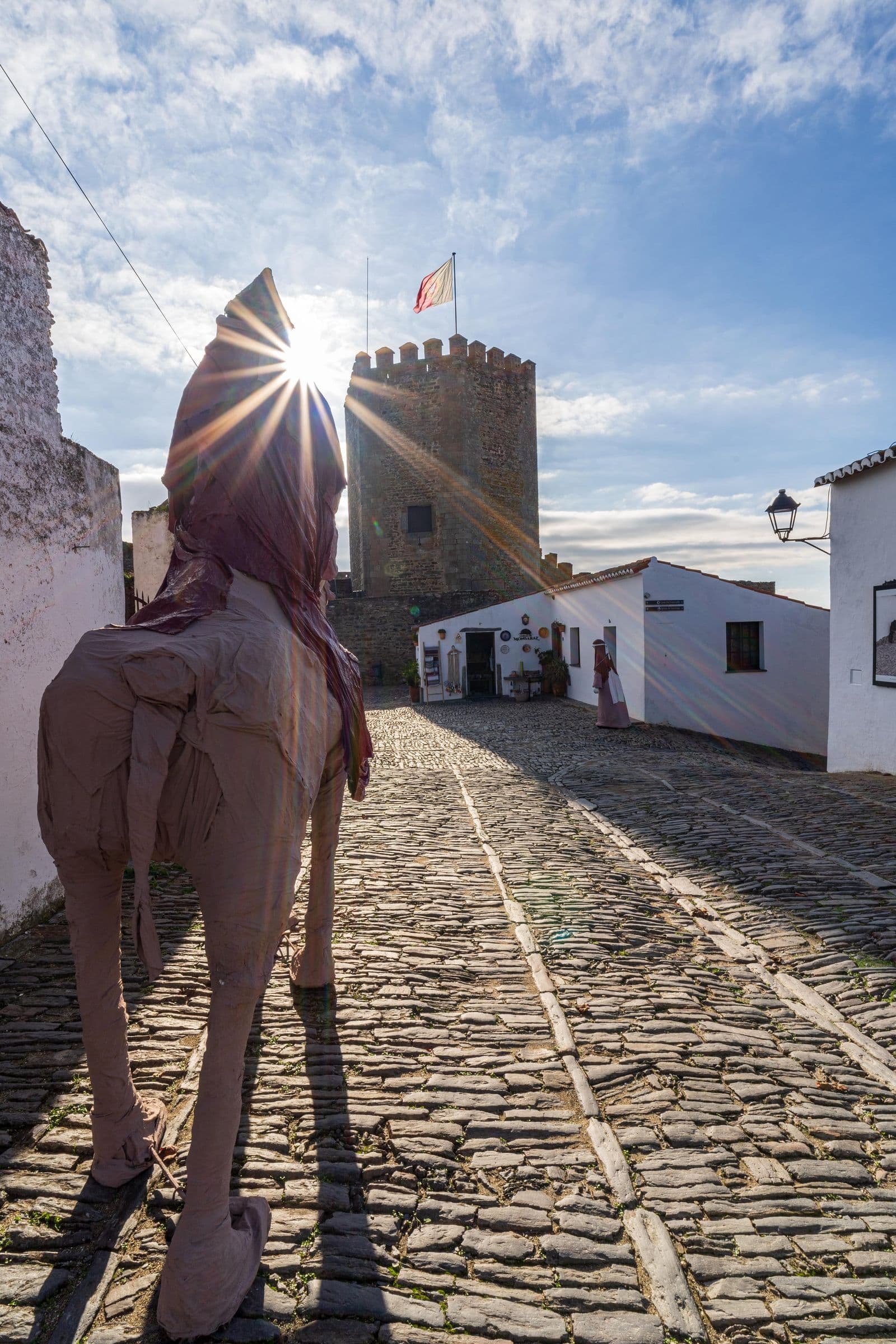 A narrow cobbled street in Monsaraz climbing towards the castle keep between whitewashed houses, with the sun low.