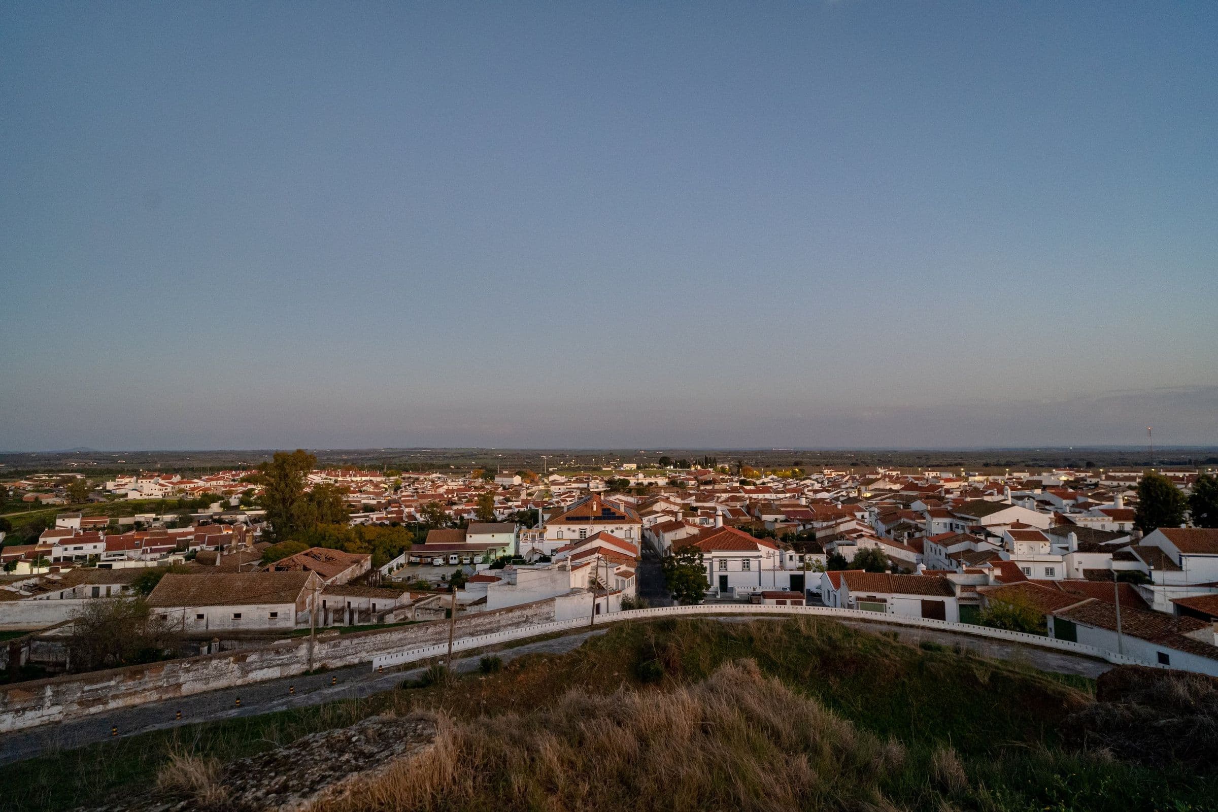 A vila de Mourão vista de cima ao fim da tarde: telhados de telha, casas caiadas e a planície a estender-se até ao horizonte.